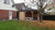 Een gezellig Interflex houten paviljoen met bank, genesteld naast een bakstenen huis, omgeven door gevallen herfstbladeren en een eenzame kale boom, bekroond met Interflex dakshingles.