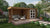 Houten tuinhuis Maja 300 x 240 x 228 cm + Luifel 340 cm van Tuindeco. Een bruin houten tuinhuis met een overdekt terras, compleet met zitmeubilair en planten, staat in een groene tuin.