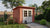 Een robuust, rood houten tuinhuis Mila (290x260x228cm) van Tuindeco, met een witgeschilderde deur. Het tuinhuis staat in een verzorgde tuin met een stenen pad, omringd door groenblijvende struiken en bomen.