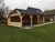 Een nieuwbouw houten Houten tuinhuis Wolfskap WKS1 met een zadeldak bedekt met dakshingles en open zijkanten op een goed onderhouden gazon, grenzend aan een woonwijk.