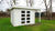 Een schilderachtig wit Interflex tuinhuis met grote ramen en een dubbele deur, voorzien van een aangebouwde pergola aan de ene kant en een zijluifel, gelegen op een weelderig groen gazon met bomen op de achtergrond.